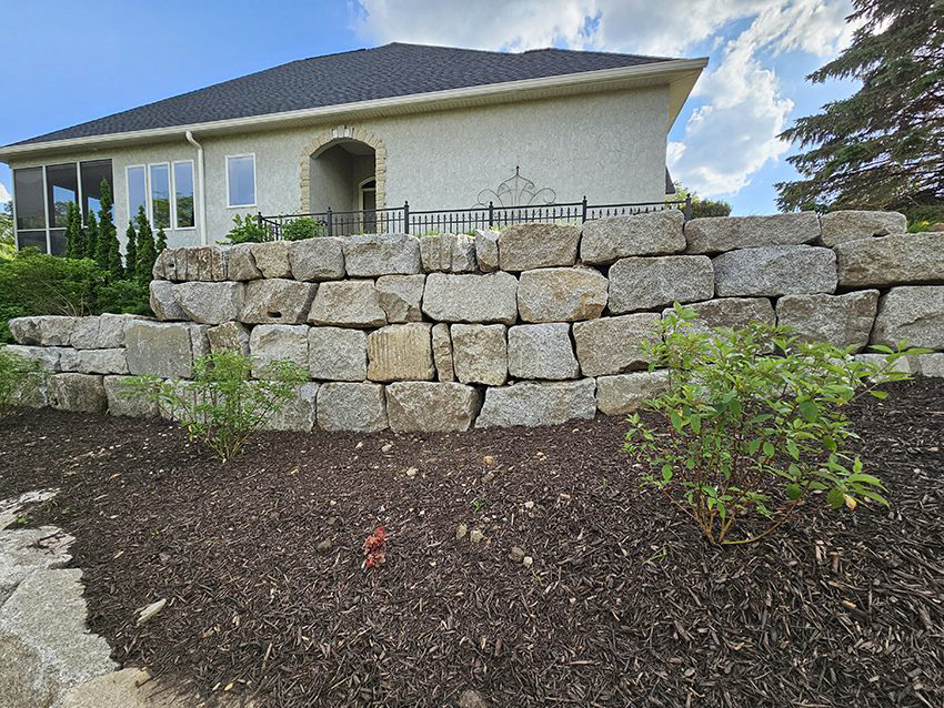 Tiered granite boulder retaining wall with mulched planting beds in front of a residential home built by Triple Landscaping